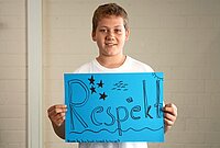 Ein Junge in weißem T-Shirt hält ein blaues Poster mit dem Wort „Respekt“ in großen, dekorativen Buchstaben. Das Poster zeigt Sterne, Wellen, Sonne und Fischmotive. Der Hintergrund besteht aus einer hellen Ziegelwand.