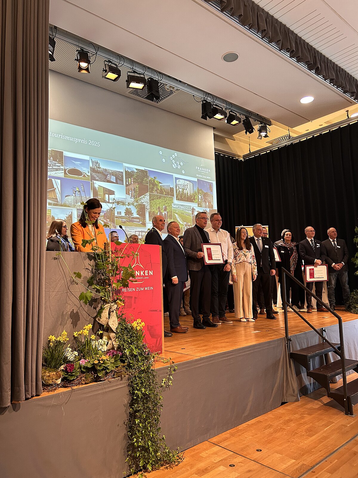 Preisverleihung auf Bühne mit rotem „WILHELMSEN“-Pult und Blumenarrangement – mehrere Personen halten gerahmte Urkunden, Hintergrund zeigt Präsentationsfolie „Wilhelmsen Sustainability Award 2023“ mit Bildern und Texten zu Nachhaltigkeit, Szene vermittelt feierliche Anerkennung für nachhaltiges Engagement in professionellem Rahmen.