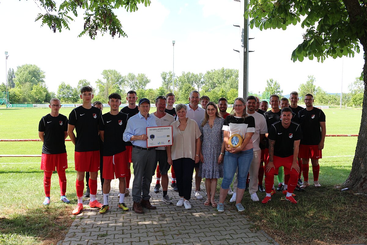 Viele Personen posieren auf einem Fußballfeld, eine Dame hat den Defibrillator in der Hand