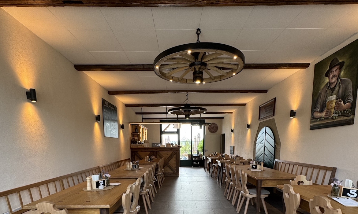 Rustikal eingerichteter Gastraum mit langen Holztischen und naturbelassenen Stühlen – die Decke zeigt freiliegende Holzbalken und zwei große Kronleuchter im Wagenradstil mit warmem Licht; helle Wände mit Wandlampen und gerahmten Bildern, darunter ein großes Gemälde mit Biermotiv; im Hintergrund Barbereich mit Flaschenregalen und großen Fenstern mit Blick ins Grüne – Atmosphäre wirkt traditionell, gemütlich und einladend.