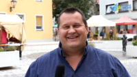 Mann mit Mikrofon steht auf einem gepflasterten Platz und lächelt in die Kamera; im Hintergrund Marktstände und Menschen bei einer Veranstaltung.