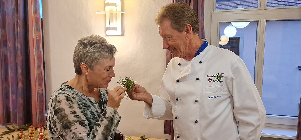 Zwei Personen stehen in einem Innenraum. Eine trägt eine weiße Kochjacke mit blauer Paspel und der Aufschrift „Medizinische Küche – G. Vollkommer“ und reicht der anderen, die ein gemustertes Oberteil trägt, frische Kräuter zum Riechen. Im Hintergrund sind ein gedeckter Tisch, violette Vorhänge und Fenster mit Lichtreflexionen sichtbar.