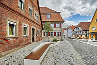 Gepflasterte Straße in einer historischen Altstadt mit Fachwerkhäusern und Gebäuden aus rotem Backstein und Sandstein, alle mit roten Ziegeldächern; im Vordergrund eine moderne Sitzbank aus Beton und Holz, daneben ein kleiner Baum; im Hintergrund weitere traditionelle Häuser, darunter ein gelbes Gebäude mit Ladeneingang, und ein teilweise bewölkter Himmel mit blauen Abschnitten.