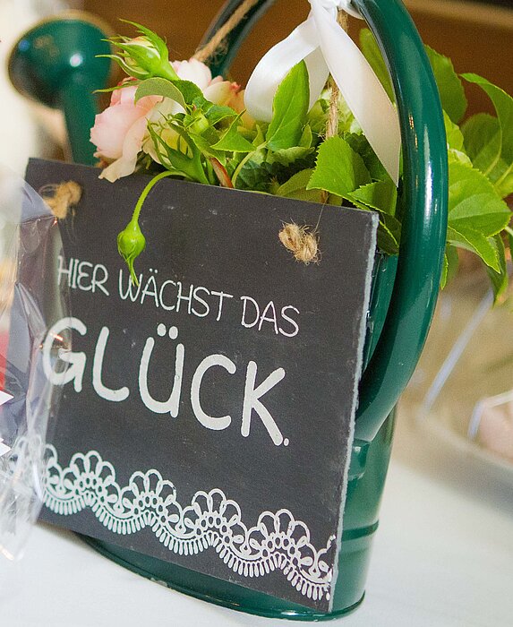 Ein grüner Metall-Blumentopf mit Griff enthält rosa Blüten und grüne Blätter, daran hängt ein schwarzes Schild mit weißer Aufschrift „HIER WÄCHST DAS GLÜCK“ und einer dekorativen Spitzenbordüre am unteren Rand.