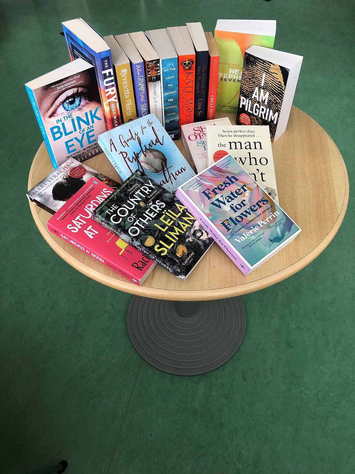 Runder Holztisch mit mehreren englischsprachigen Büchern in unterschiedlichen Farben und Formaten, darunter Titel wie „In the Blink of an Eye“, „I Am Pilgrim“, „Fresh Water for Flowers“, „The Country of Others“ und „Saturdays at Noon“, auf grünem Boden.