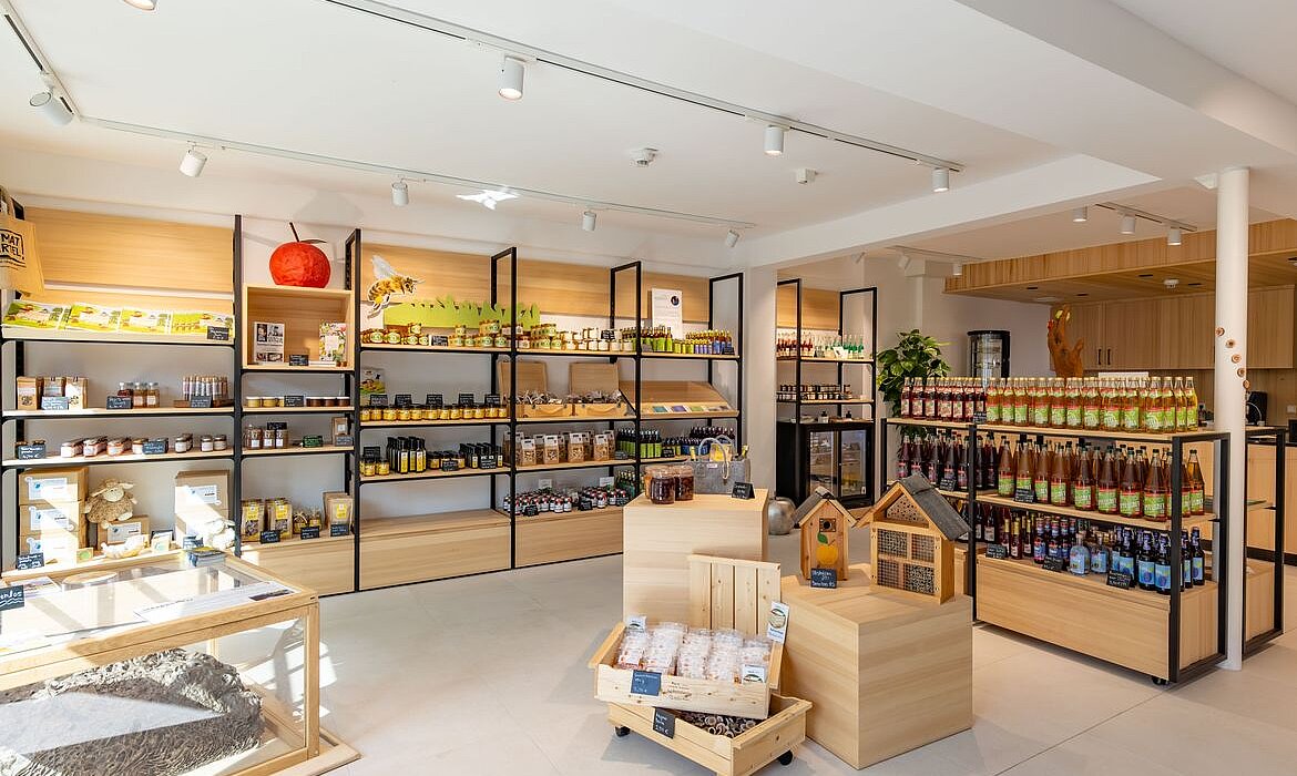 Innenraum eines Naturwarenladens mit Holzregalen und zentralem Präsentationstisch – Produkte wie Honiggläser, Getränke, verpackte Lebensmittel und ein Modell eines Bienenhauses deuten auf Schwerpunkt Imkerei und regionale Erzeugnisse; helle Beleuchtung und Pflanzen schaffen ein einladendes, organisch gestaltetes Ambiente.