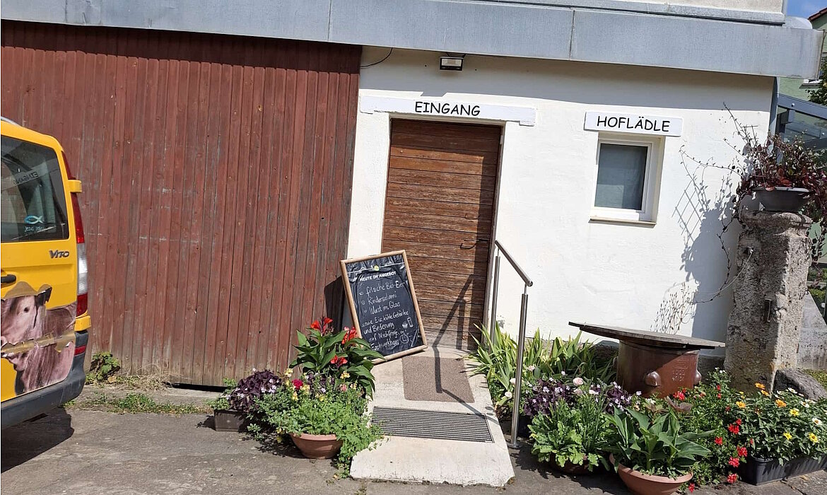 Einladender Hofladen-Eingang mit handgeschriebener Tafel: Frisches Fleisch vom Weiderind und Schwein, Grillpakete, Wurst, Käse, Eier, Milch, Joghurt, Eingemachtes, Nudeln, Marmelade, Honig, Säfte und Wein – ein echtes Schaufenster regionaler Vielfalt und bäuerlicher Qualität.