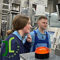 Zwei Personen in blauer Arbeitskleidung stehen vor einem Maschinenbedienfeld mit Monitoren und technischen Unterlagen – Szene in einem Industrie- oder Ausbildungsumfeld mit Bezug zur „JOB JUNGLE“-Initiative und zur Firma KRONES.