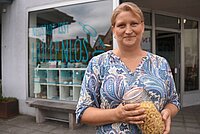 Frau mit blau-weiß gemustertem Oberteil steht lächelnd vor einem Geschäft und hält ein Glas mit Pasta – hinter ihr ein Fenster mit grafischen Elementen und dem Schriftzug „WENIGER IST LOST HOLFLNLOS“, Szene deutet auf nachhaltiges Einkaufen und bewusstes Konsumverhalten hin.