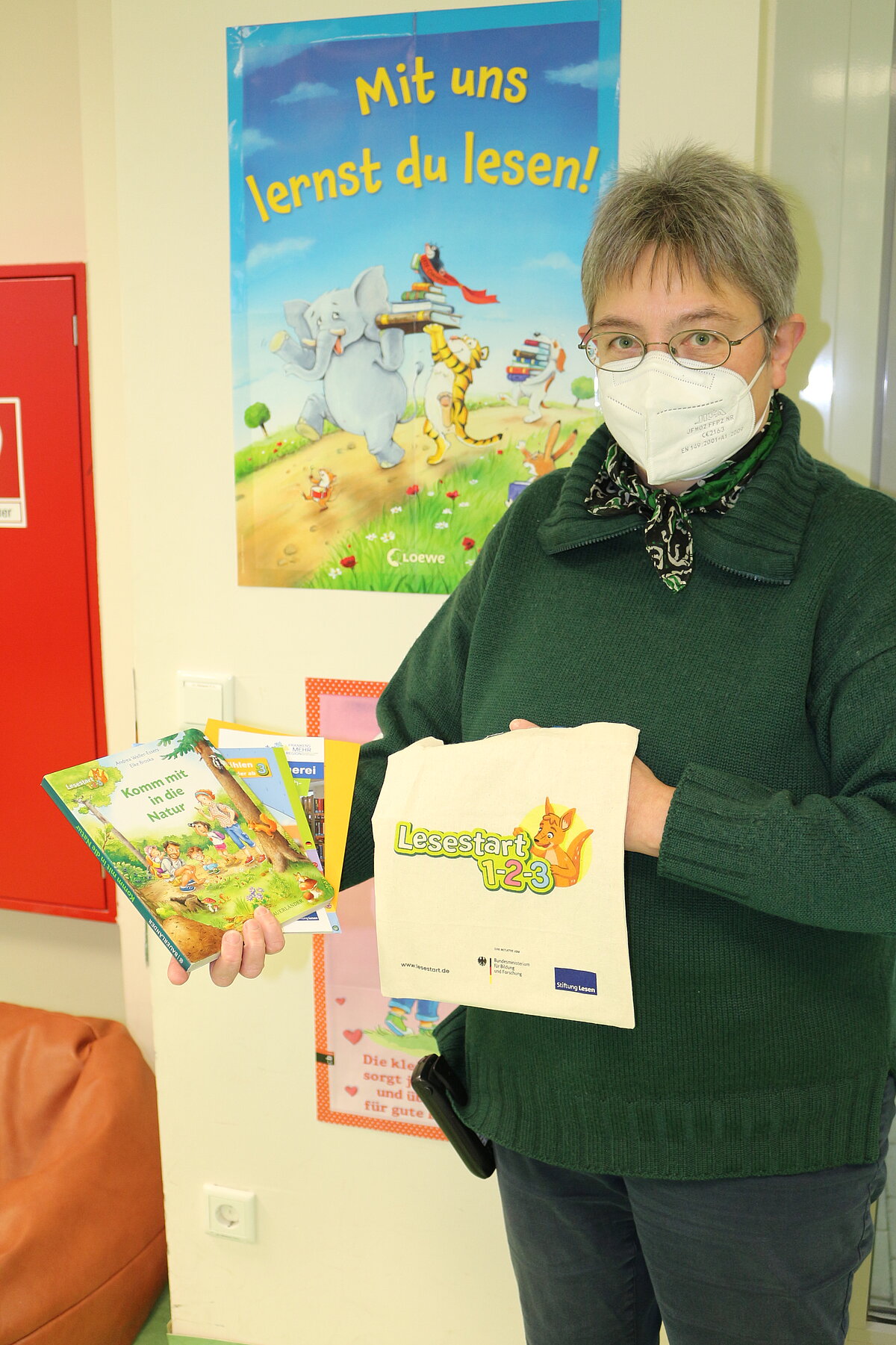 Eine Person mit Brille, grünem Pullover und weißer Maske hält zwei Kinderbücher und eine Stofftasche mit der Aufschrift „Lesestart 1-2-3“ sowie einem Fuchs-Logo. Im Hintergrund hängen bunte Poster mit Lesemotiven, darunter eines mit dem Text „Mit uns lernst du lesen!“.