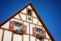 Ein charmantes Stück fränkischer Baukunst: Das Fachwerkhaus mit seinen roten Fensterläden, blühenden Blumenkästen und kunstvoll verzierten Balken strahlt im klaren Blau des Himmels – ein liebevoll gepflegtes Detail, das Geschichte und Heimatgefühl vereint.