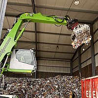 Große Industriehalle mit SENNEBOGEN-Greifarm, der einen Metallkorb voller gepresstem Papier und Karton anhebt – im Hintergrund Papierberge, roter Container und eine Person in gelbem Shirt bei der Abfallverarbeitung.