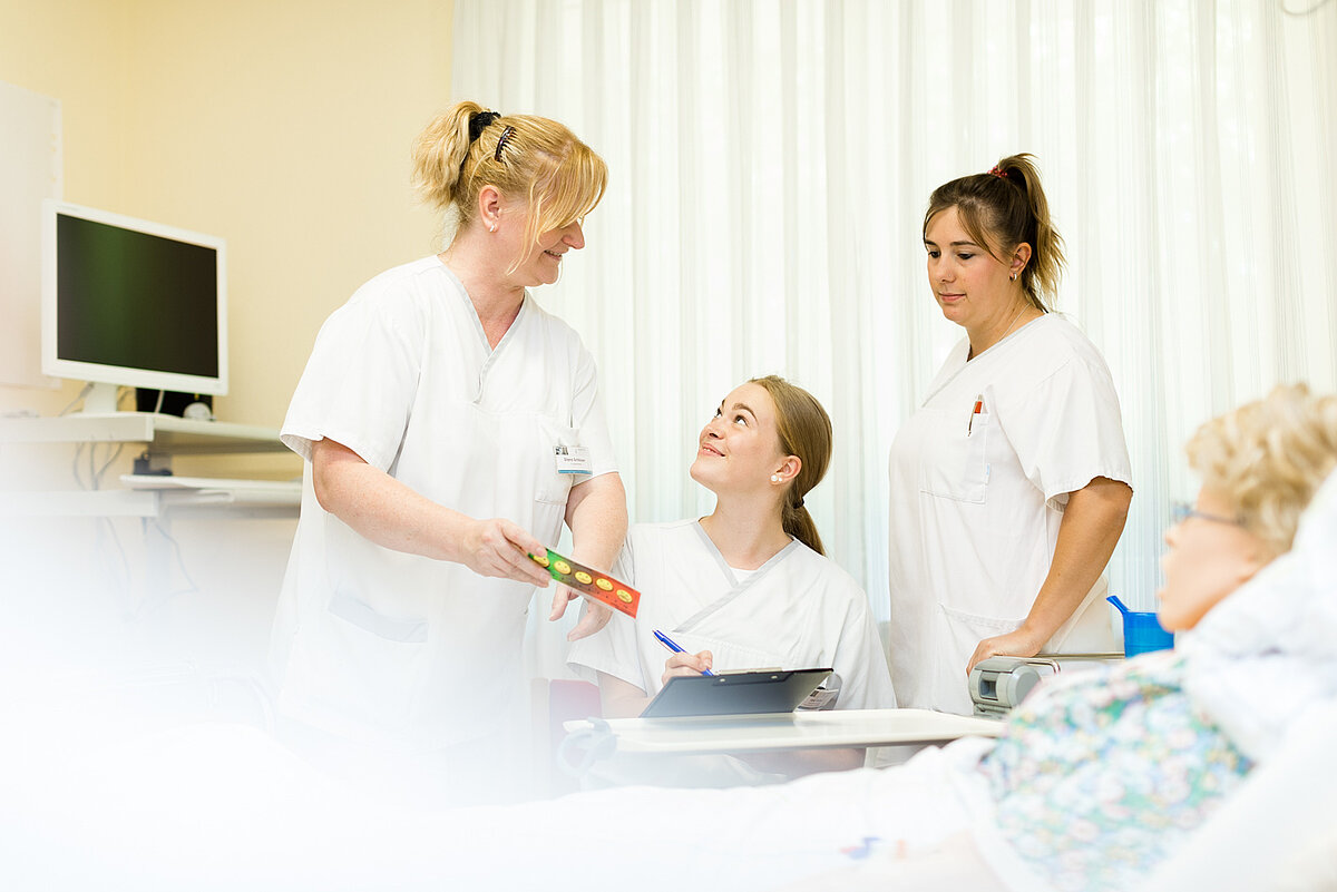 Vier Personen in einem Klinikzimmer, drei davon in weißer Berufskleidung. Eine Person liegt im Bett, zwei stehen, eine sitzt und lächelt, während sie ein Farbschema-Tool entgegennimmt. Im Hintergrund sind medizinische Geräte und Wandtelefon sichtbar.