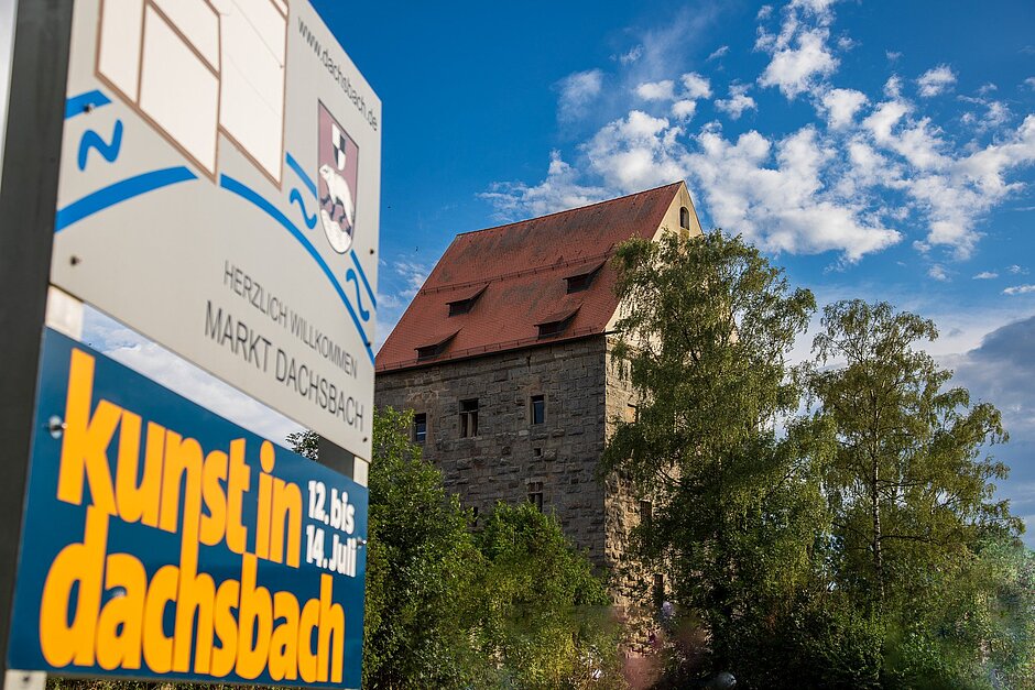 Roll-up mit dem Veranstaltungshinweis und dem Wasserschloss Dachsbachi m Hitnergrund, ein hohes steineres Gebäude mit roten Ziegeln 