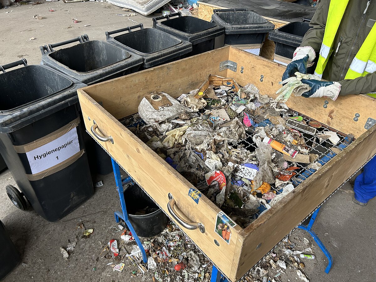 Ein Sortiertisch mit verschiedenen Abfällen wie Papier, Plastik und Verpackungen steht in einer Halle, daneben mehrere schwarze Mülltonnen mit Beschriftung „Hygienepapier“. Eine Person trägt eine Warnweste und Handschuhe und hält Abfallstücke über dem Tisch.