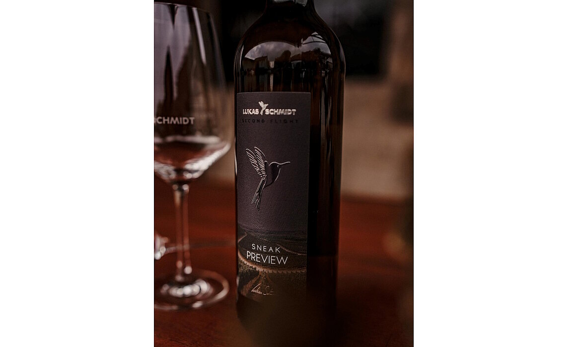 Nahaufnahme einer Weinflasche mit dunklem Etikett „LUKAS SCHMIDT SECOND FLIGHT“ und Kolibri-Illustration – darunter Schriftzug „SNEAK PREVIEW“, daneben passendes Weinglas mit „SCHMIDT“-Gravur, beide auf Holzoberfläche vor unscharfem Hintergrund arrangiert, Szene vermittelt hochwertige Markenästhetik und exklusive Produktpräsentation.