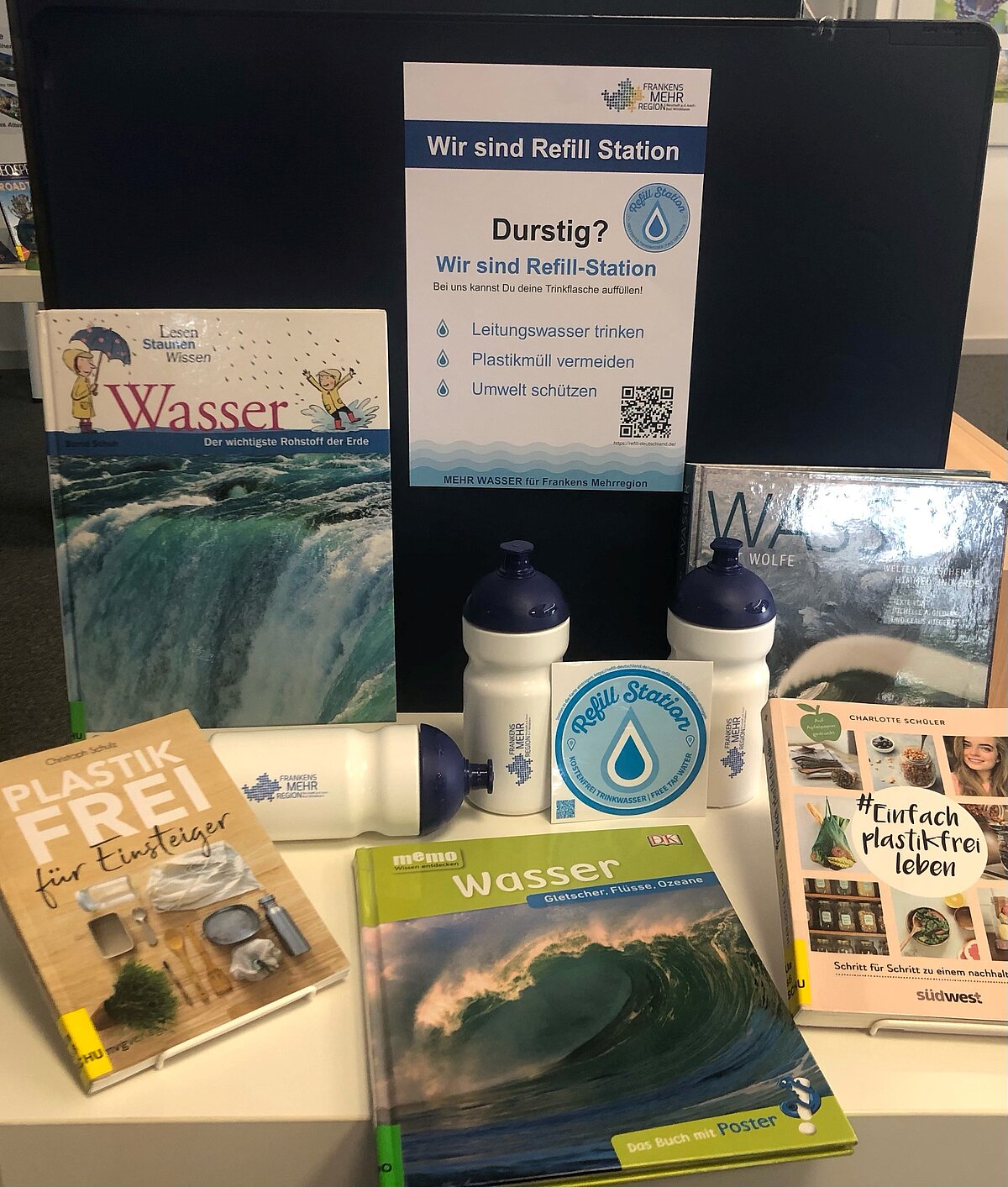 Ein Tischdisplay mit schwarzem Hintergrund zeigt Bücher zum Thema Wasser und Plastikvermeidung, mehrere wiederverwendbare Trinkflaschen mit „Refill Station“-Aufdruck sowie ein Schild mit dem Hinweis, dass hier kostenlos Leitungswasser aufgefüllt werden kann. Ein QR-Code und Informationsmaterial sind ebenfalls sichtbar.
