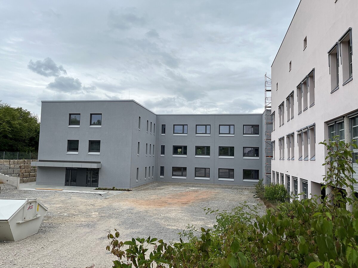 Neubau eines grauen, dreistöckigen Gebäudes mit mehreren Fenstern und Kiesfläche im Vordergrund, rechts angrenzend ein älteres weißes Gebäude, im Hintergrund Bäume und bewölkter Himmel.