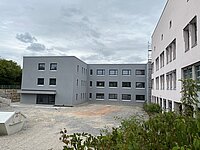 Neubau eines grauen, dreistöckigen Gebäudes mit mehreren Fenstern und Kiesfläche im Vordergrund, rechts angrenzend ein älteres weißes Gebäude, im Hintergrund Bäume und bewölkter Himmel.