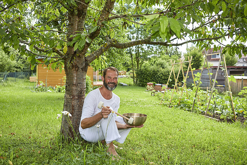 Armin Nögel in weißem Shirt und Hose mt einer Klangschale in Gold unter einem Baum mit Wiese im Hintrgrund