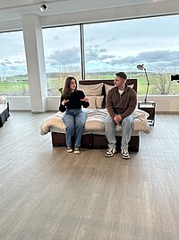 Zwei Personen sitzen auf einem Bett in einem modern eingerichteten Raum mit großen Fenstern und sprechen miteinander; die Einrichtung ist hell und schlicht, mit Blick ins Grüne.