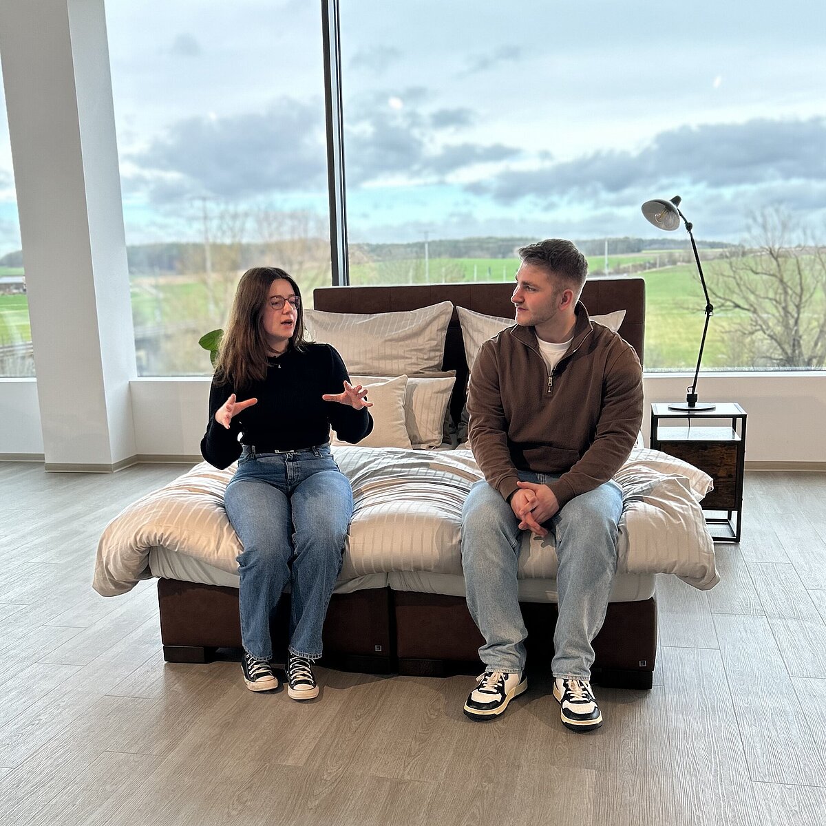 Zwei Personen sitzen auf einem Bett in einem modern eingerichteten Raum mit großen Fenstern und sprechen miteinander; die Einrichtung ist hell und schlicht, mit Blick ins Grüne.