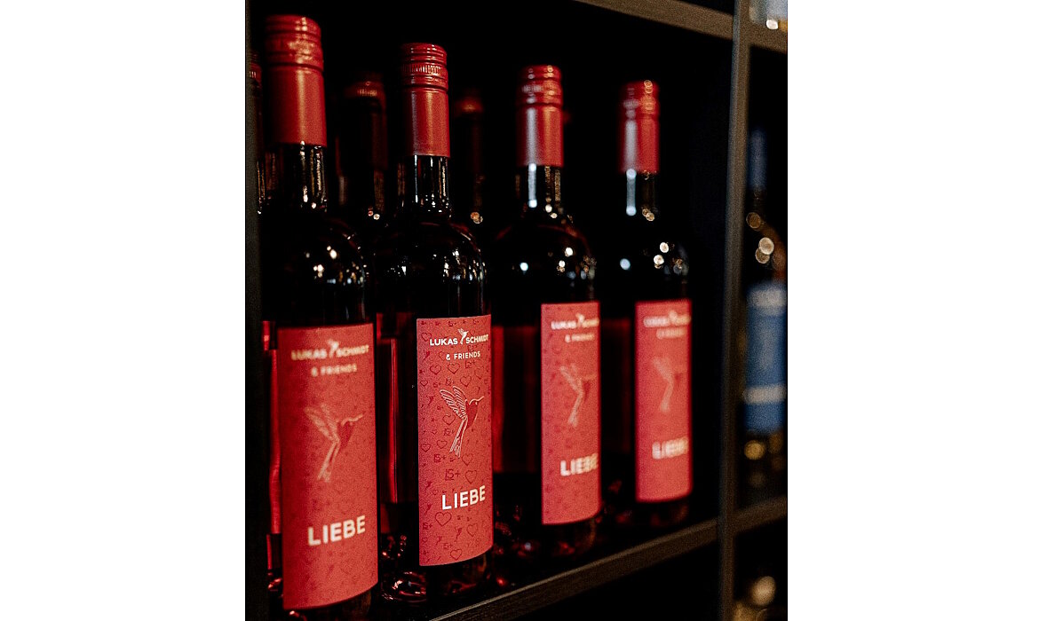 Weinregal mit mehreren Flaschen Rotwein der Marke „LIEBE“ – Flaschen mit roten Etiketten und Schraubverschlüssen, Aufdruck „LAURA ZIEGLER & FRIENDS“ sowie stilisierte Figur in Bewegung, Flaschen ordentlich in Reihen angeordnet, daneben Regal mit blauen Etiketten, Szene zeigt markantes Verpackungsdesign und strukturierte Präsentation im Einzelhandel.