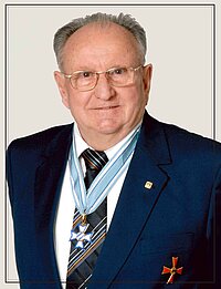 Eine Person trägt einen dunkelblauen Anzug mit weißem Hemd und gestreifter Krawatte. Um den Hals hängt ein hellblaues Band mit einem Orden in Form eines Kreuzes, und am linken Revers ist ein kleiner Anstecker befestigt. Auf der linken Brustseite des Anzugs befindet sich ein weiteres Abzeichen in Form eines roten Kreuzes.