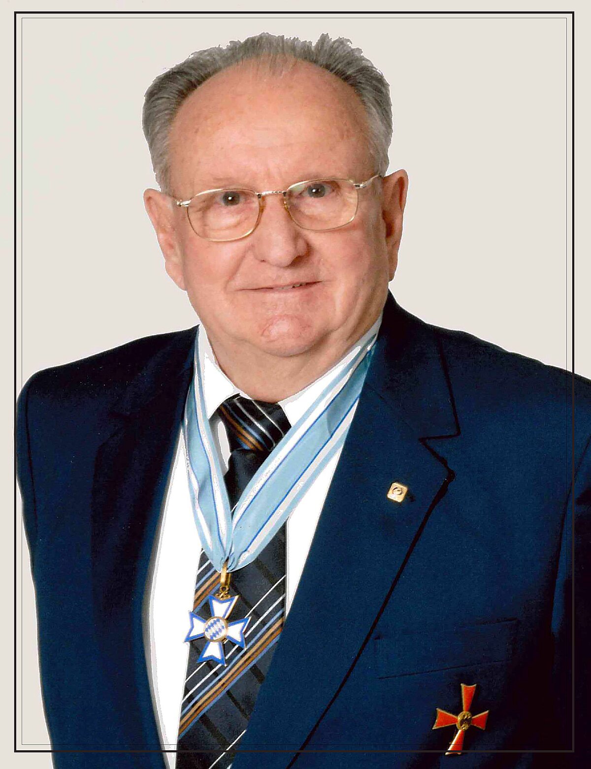 Eine Person trägt einen dunkelblauen Anzug mit weißem Hemd und gestreifter Krawatte. Um den Hals hängt ein hellblaues Band mit einem Orden in Form eines Kreuzes, und am linken Revers ist ein kleiner Anstecker befestigt. Auf der linken Brustseite des Anzugs befindet sich ein weiteres Abzeichen in Form eines roten Kreuzes.