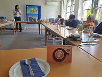 Konferenzraum mit U-förmig angeordneten Holztischen, auf denen Teller, Besteck, Gläser und Getränkeflaschen stehen, im Vordergrund ein Teller mit Serviette und Löffel sowie eine Karte mit dem Text „Vielfalt hat viele schöne Ansichten und Seiten“ und einem Bild von Gebäck; im Hintergrund eine Person steht vor einem Tisch mit weiteren Getränken und Lebensmitteln, an der Wand hängt ein buntes Poster mit einer Ernährungspyramide.