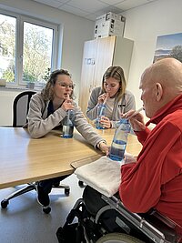 Drei Personen sitzen an einem Holztisch in einem Pflege- oder Betreuungskontext und trinken mit langen Strohhalmen aus Wasserflaschen; eine ältere Person im Rollstuhl trägt einen roten Pullover, zwei Betreuungskräfte in grauen Jacken begleiten die Szene.