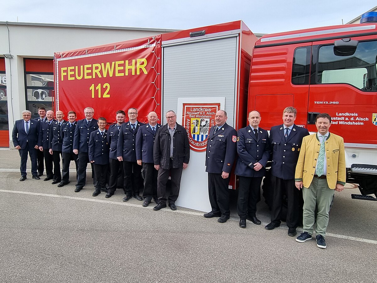 Gruppe in Uniform und Zivilkleidung posiert vor einem Feuerwehrfahrzeug mit der Aufschrift „FEUERWEHR 112“ – Aufnahme vermutlich bei einer offiziellen Veranstaltung oder Übergabe vor dem Feuerwehrhaus.