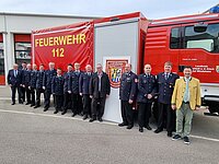 Gruppe in Uniform und Zivilkleidung posiert vor einem Feuerwehrfahrzeug mit der Aufschrift „FEUERWEHR 112“ – Aufnahme vermutlich bei einer offiziellen Veranstaltung oder Übergabe vor dem Feuerwehrhaus.
