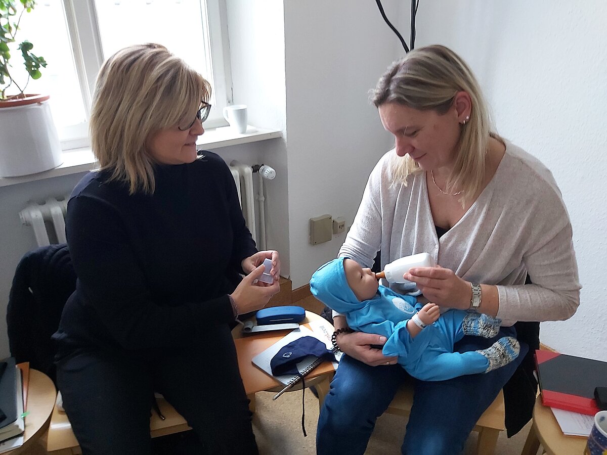Zwei Personen sitzen in einem Raum auf Holzstühlen, eine Person hält eine Babypuppe in blauer Kleidung und gibt ihr mit einer weißen Flasche zu trinken, die andere Person hält ein kleines Behältnis in der Hand. Auf dem Tisch liegen verschiedene Utensilien wie eine Tasche und ein Notizbuch, im Hintergrund ein Fenster mit weißer Fensterbank und einem Becher darauf.