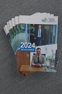 Mehrere Broschüren liegen fächerförmig auf grauem Stoff, Titel „2024 Jahresrückblick“ mit dem Logo „Frankens MEHR Region Neustadt a.d.Aisch-Bad Windsheim“, auf dem Cover ein Foto von einer Person im Anzug an einem Schreibtisch und ein weiteres Foto von einer Person im Eingangsbereich eines Gebäudes.