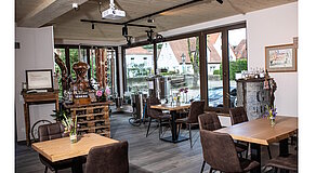 Gemütlicher Gastraum mit Holzdecke und -boden, hellen Tischen und gepolsterten Stühlen – Kupferdestille auf Holzpodest, Blumenvasen auf den Tischen, große Fenster mit Blick auf eine ruhige Straße; rustikale Details und moderne Ausstattung schaffen ein einladendes Ambiente.