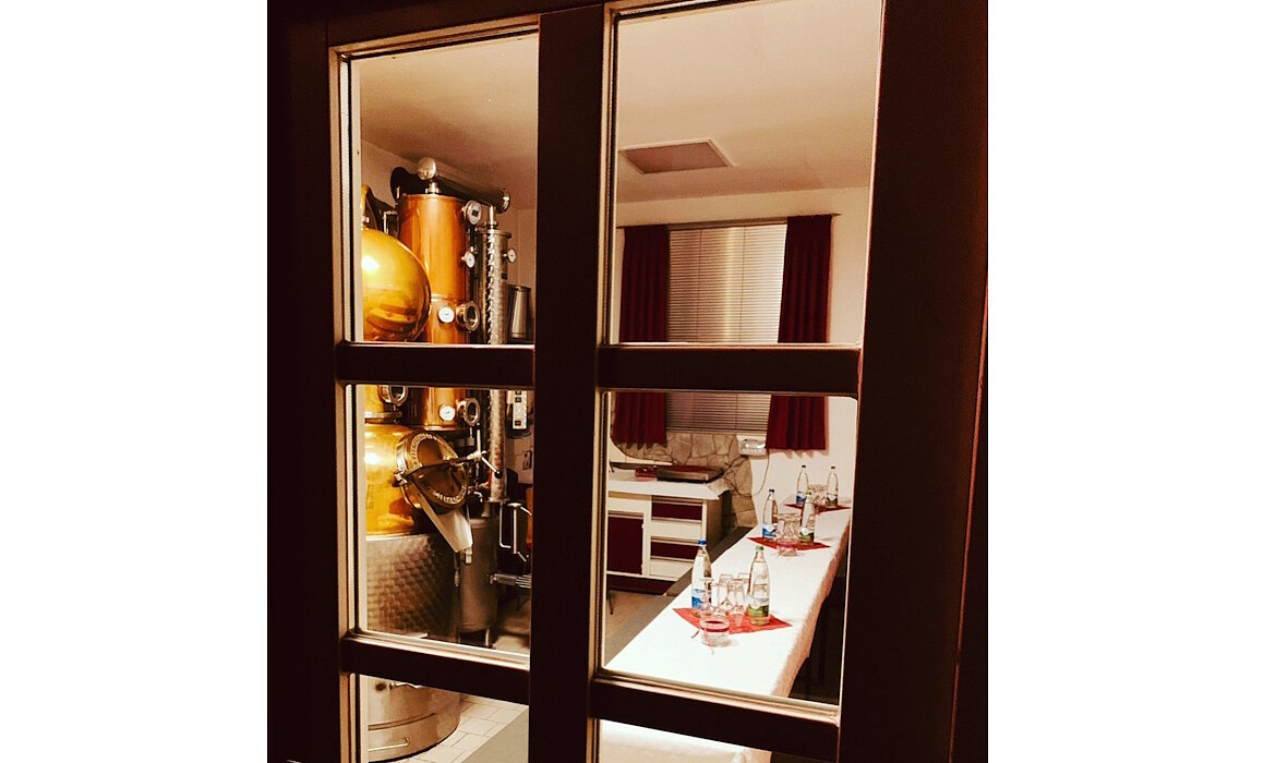 Ein Blick durch die Glastür offenbart eine stimmungsvolle Brennerei mit glänzender Kupferanlage und einem liebevoll gedeckten Tisch – ein Ort, an dem Handwerk und Genuss aufeinandertreffen. Die roten Vorhänge und das warme Licht verleihen dem Raum eine einladende Atmosphäre.