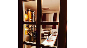 Ein Blick durch die Glastür offenbart eine stimmungsvolle Brennerei mit glänzender Kupferanlage und einem liebevoll gedeckten Tisch – ein Ort, an dem Handwerk und Genuss aufeinandertreffen. Die roten Vorhänge und das warme Licht verleihen dem Raum eine einladende Atmosphäre.