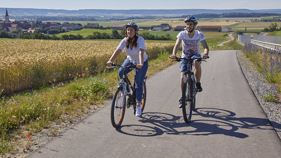 Zwei Radfahrer radeln auf einem Radweg entlang eines Getreidefeldes mit wunderschönem Weitblick.
