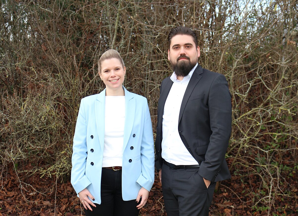 Zwei Personen stehen im Freien vor dichtem Buschwerk, beide tragen formelle Kleidung: eine Person einen hellblauen Blazer über weißem Oberteil und dunkler Hose, die andere einen dunklen Anzug mit weißem Hemd. Der Boden ist mit braunen Blättern bedeckt.