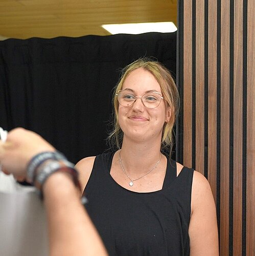 Zwei Personen stehen an einem Ladentresen; eine überreicht der anderen eine Einkaufstasche – im Hintergrund Kinderkleidung an einem Ständer, eine Pflanze und eine Holzvertäfelung mit Vorhang.