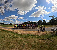 Eine Gruppe von Radfharern neben einem Radschild und Rädern vor einem bewölkten Himmel