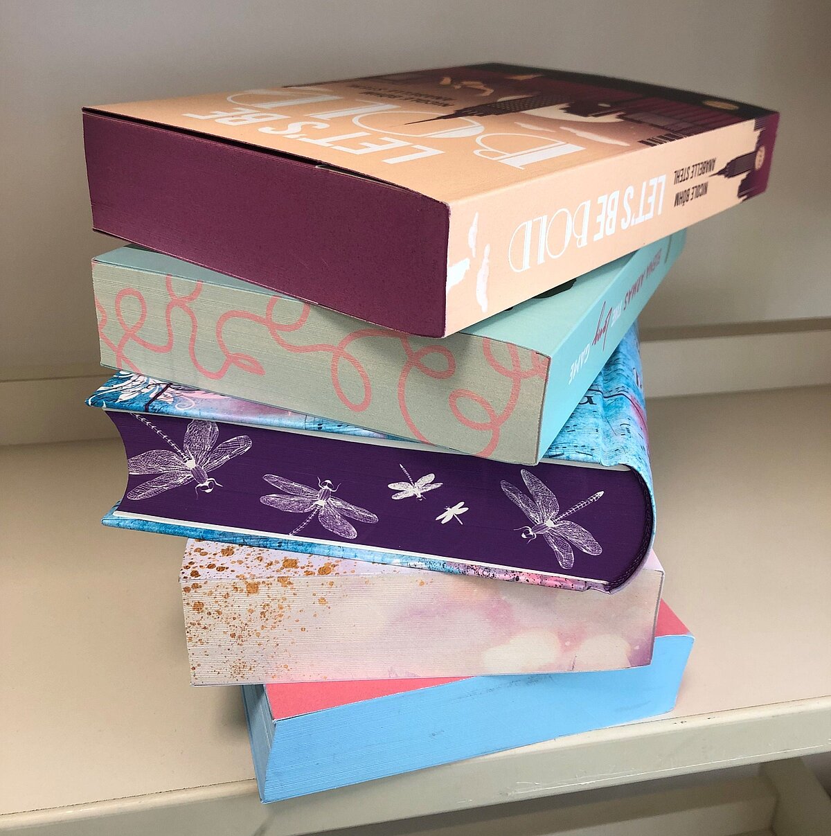 Fünf Bücher liegen gestapelt aufeinander, alle mit farbig gestalteten Buchschnitten: oben ein Buch mit dem Titel „LET’S BE BOLD“ in Beige und Dunkelrot, darunter Bücher in Pastellfarben mit Mustern wie geschwungenen Linien, Libellen und Farbverläufen in Blau, Rosa und Gold.