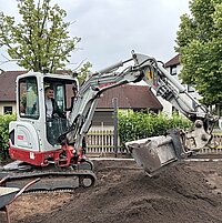 Ein Mann bedient einen Kompaktbagger auf einem Grundstück mit Häusern und Bäumen; im Vordergrund steht eine mit Erde gefüllte Schubkarre.