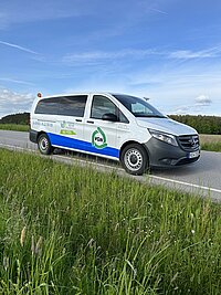 Ein weißer Mercedes-Benz Kleinbus mit blauer und grüner Beschriftung steht auf einer Landstraße neben einer grünen Wiese, auf der Fahrzeugseite sind die Logos „VGN“ und „Frankens MEHR Region“ sowie eine Telefonnummer sichtbar, im Hintergrund Bäume und blauer Himmel mit Wolken.