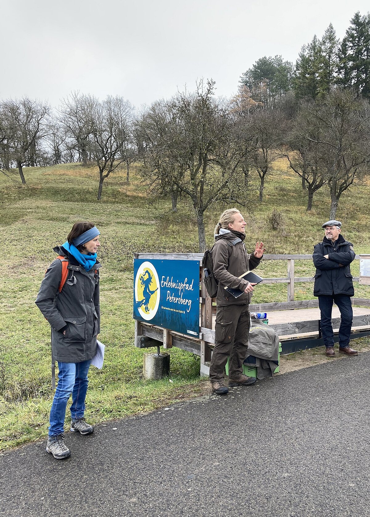 Eröffnung des barrierarmen Erlebnispfades in Marktbergel © Landratsamt Neustadt a.d.Aisch-Bad Windsheim Drei Personen an Holzplattform mit Schild „Erlebnispfad Petersberg“ – eine Person spricht gestikulierend mit Unterlagen in der Hand, zwei weitere hören aufmerksam zu, Szene zeigt Vermittlung von Natur- oder Regionalwissen in ländlicher Umgebung mit Obstbaumhang und bewölktem Himmel.