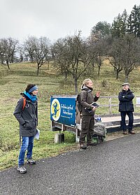Drei Personen an Holzplattform mit Schild „Erlebnispfad Petersberg“ – eine Person spricht gestikulierend mit Unterlagen in der Hand, zwei weitere hören aufmerksam zu, Szene zeigt Vermittlung von Natur- oder Regionalwissen in ländlicher Umgebung mit Obstbaumhang und bewölktem Himmel.