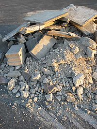 Ein Haufen aus zerbrochenem Beton mit großen Platten und kleineren Bruchstücken liegt auf dunklem Asphalt, vermutlich an einer Baustelle oder einem Abrissort.