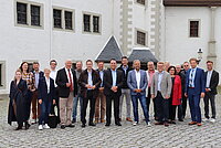 Gruppe von Personen steht auf einem gepflasterten Platz vor einem historischen Gebäude mit weißer Fassade, grauen Schieferdächern und dekorativen Steinrahmen um Türen und Fenster; im Hintergrund sind eine Laterne und architektonische Details sichtbar.