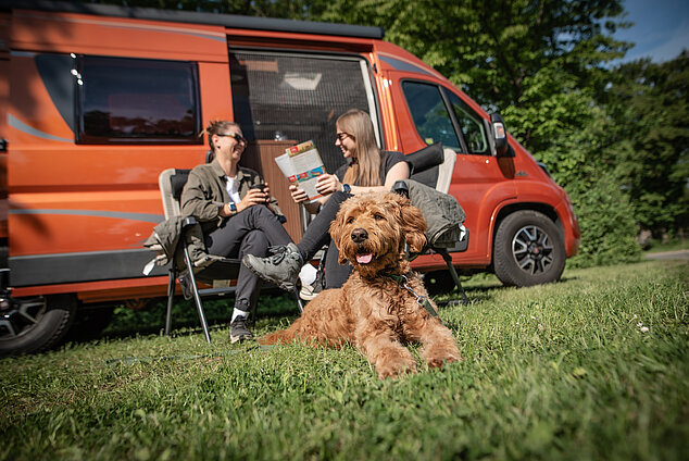 Zwei Personen und ein Hund sitzend vor einem organgefarbenen Camper 